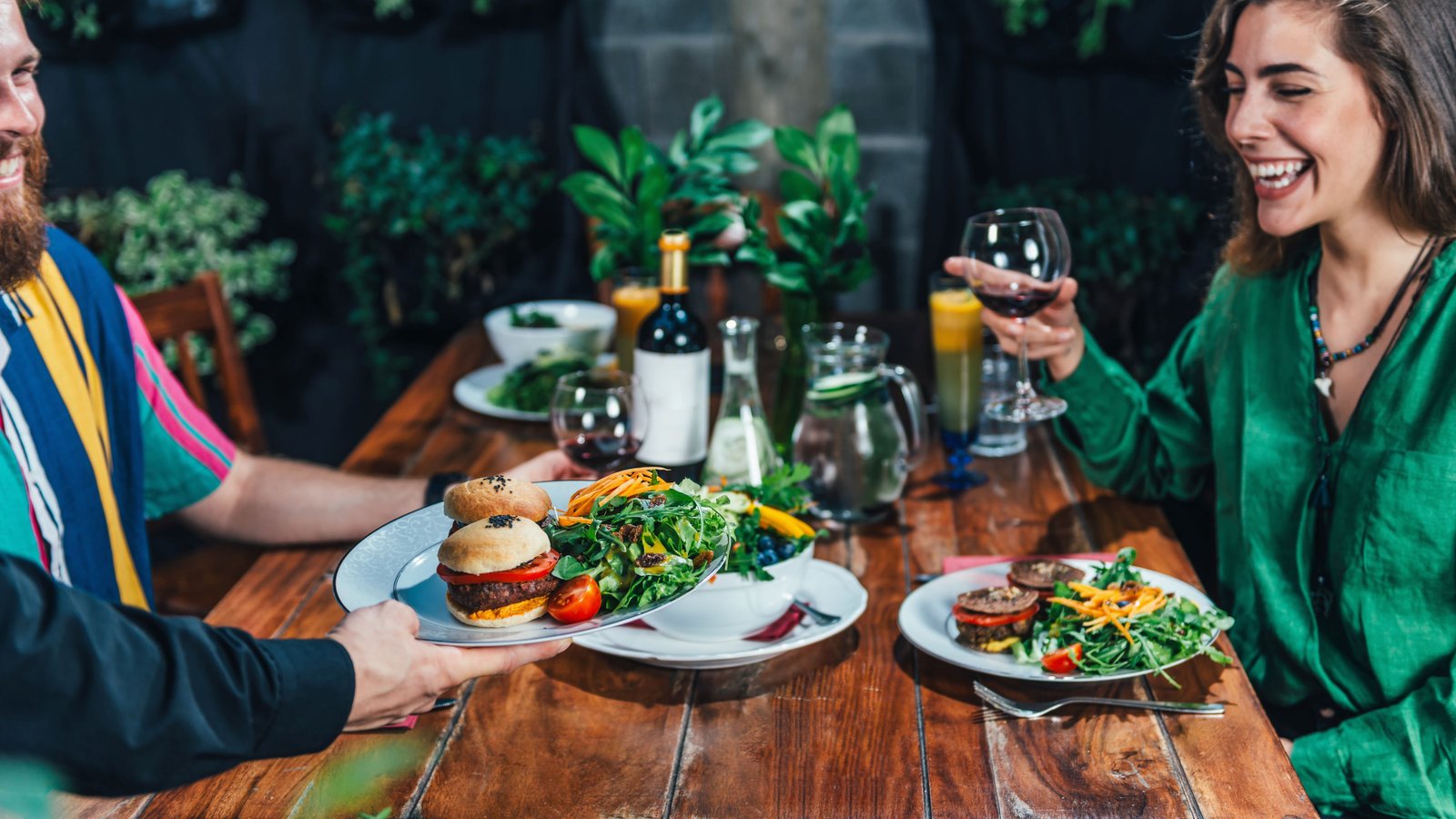 Husband and wife having their own favourite vegan and meat burger enjoying it with a glass of red wi